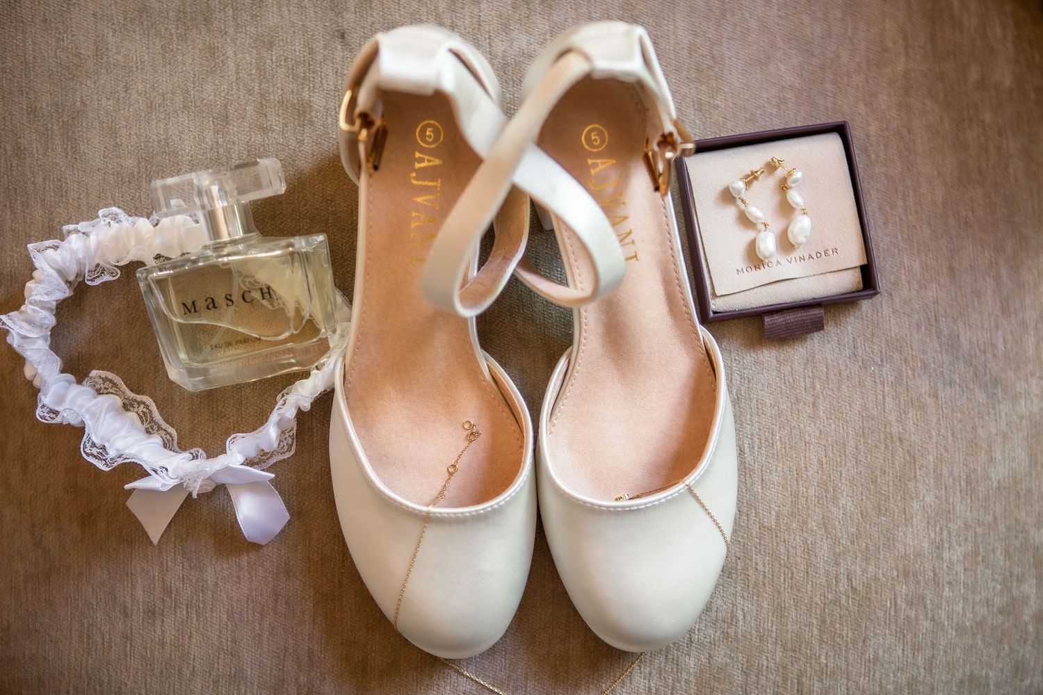 Bridal accessories including white shoes, garter, and jewelry arranged on neutral fabric.