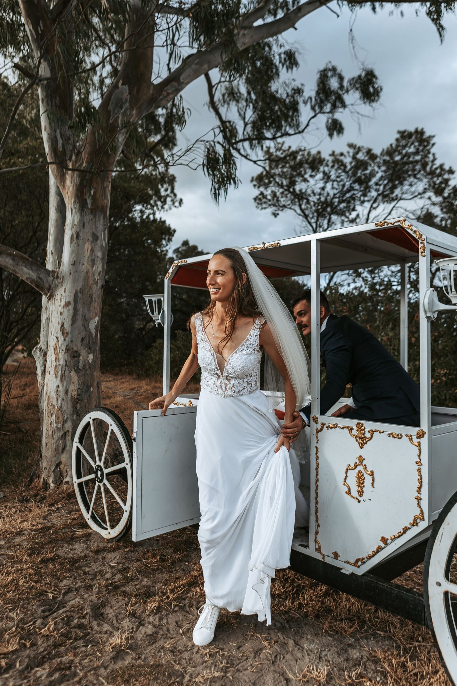 Bride in flowing white gown poses near decorative white carriage in outdoor setting.