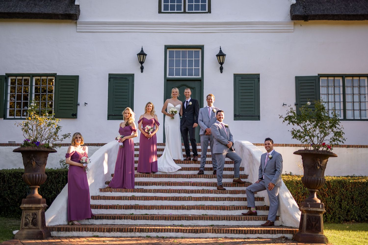 Wedding party poses on grand exterior steps of a white mansion with green shutters and formal landscaping.
