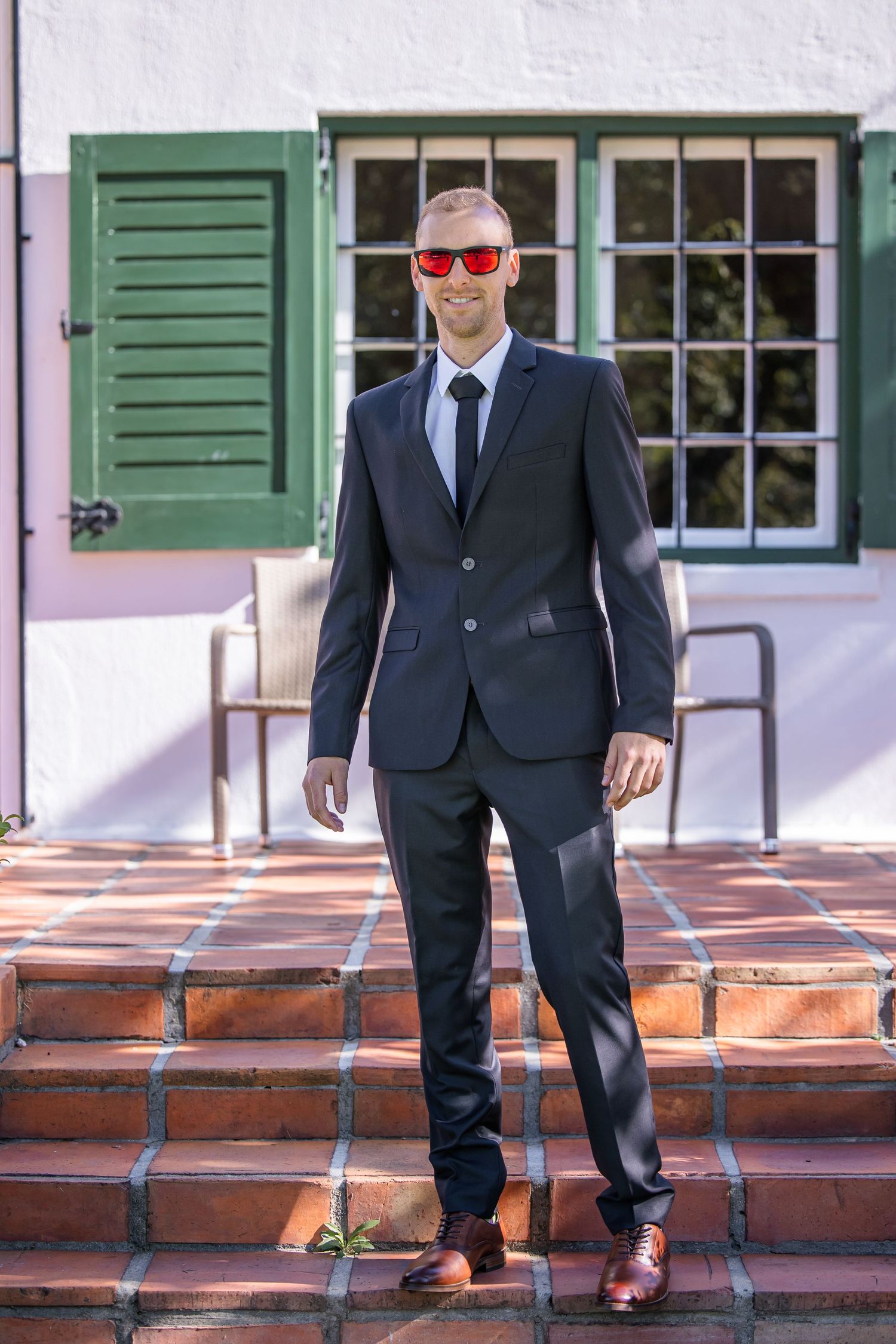 Groomsmen in dark suits standing on brick steps with green shuttered windows behind them.