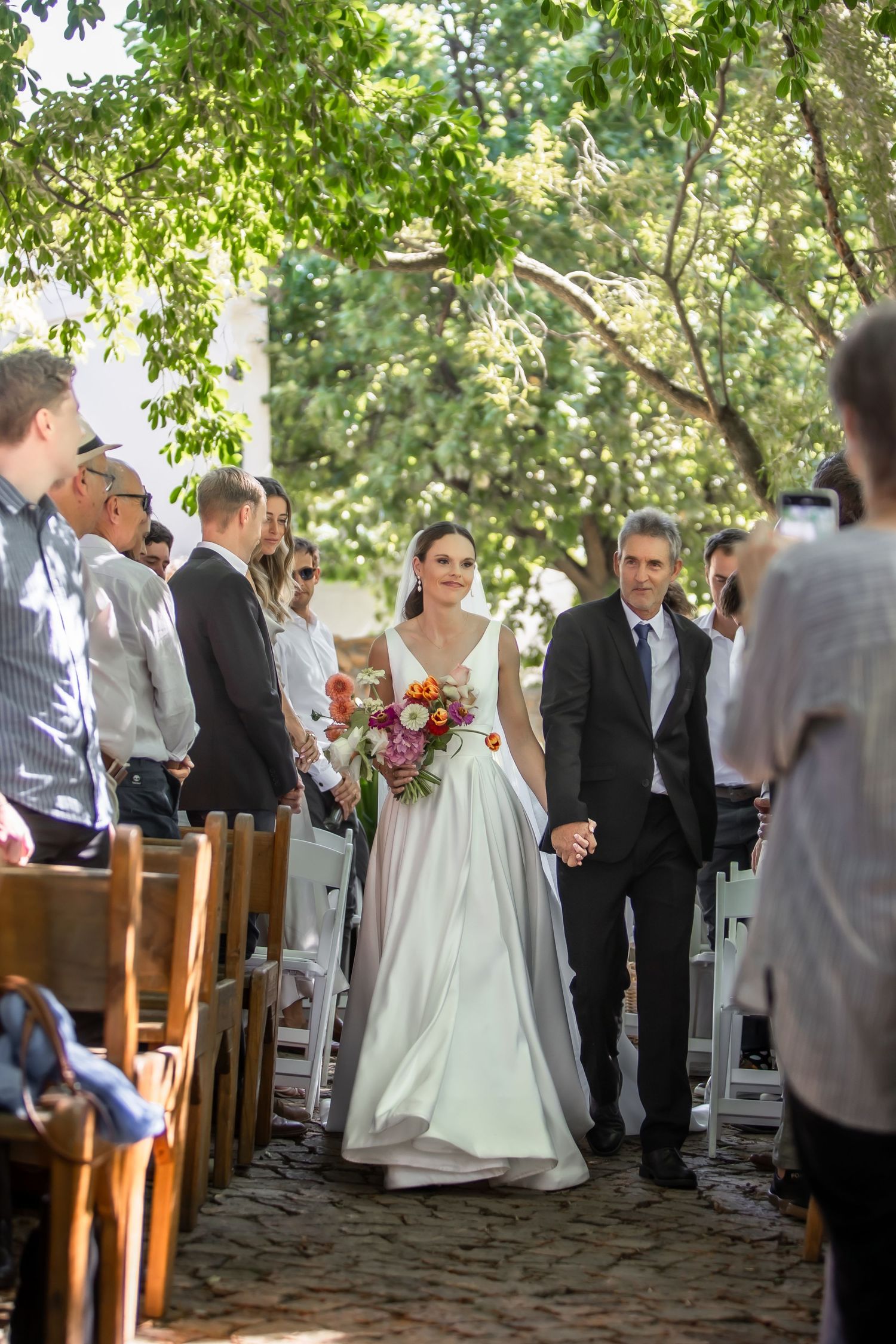 Outdoor wedding ceremony under tree canopy with wooden chairs and gathered guests.