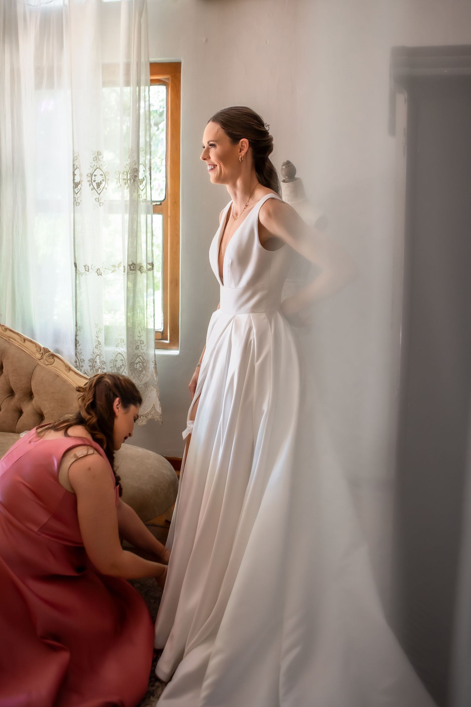 Wedding dress being fitted in a sunlit room with white curtains.