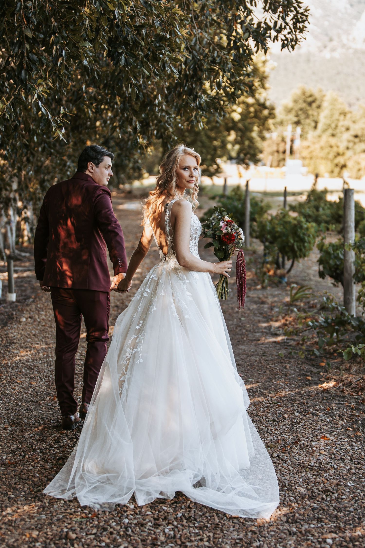 A wedding couple walks together on a leaf-covered path surrounded by trees.