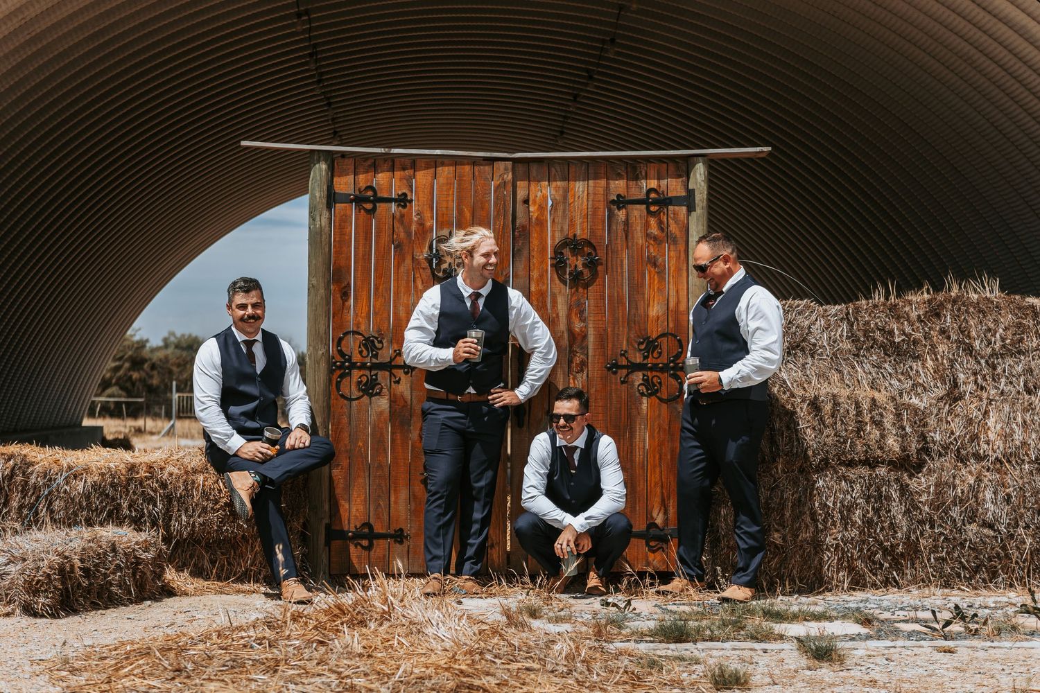 Wedding party members gather outside wooden barn door under curved metal structure.