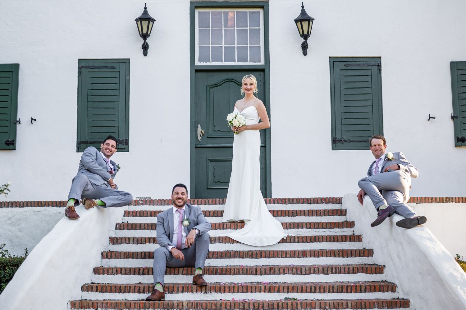 Wedding party posing playfully on white steps with green shutters and lanterns.