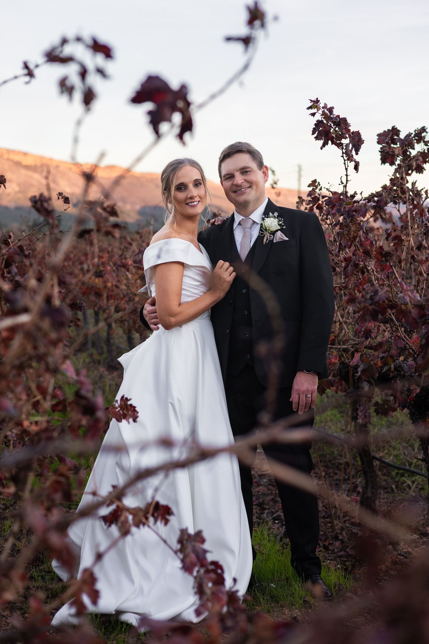 Bride and groom posing romantically among autumn vineyard vines at sunset.