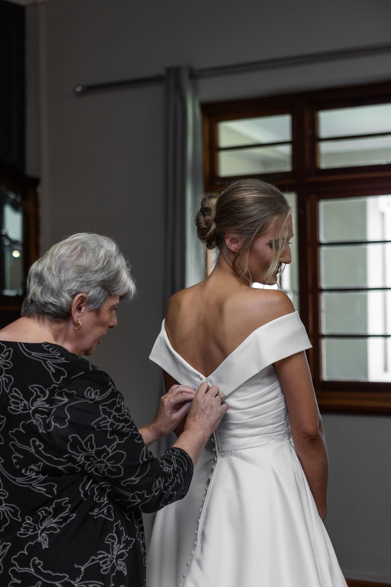 Older woman helping bride button up elegant off-shoulder white wedding gown from behind near window.