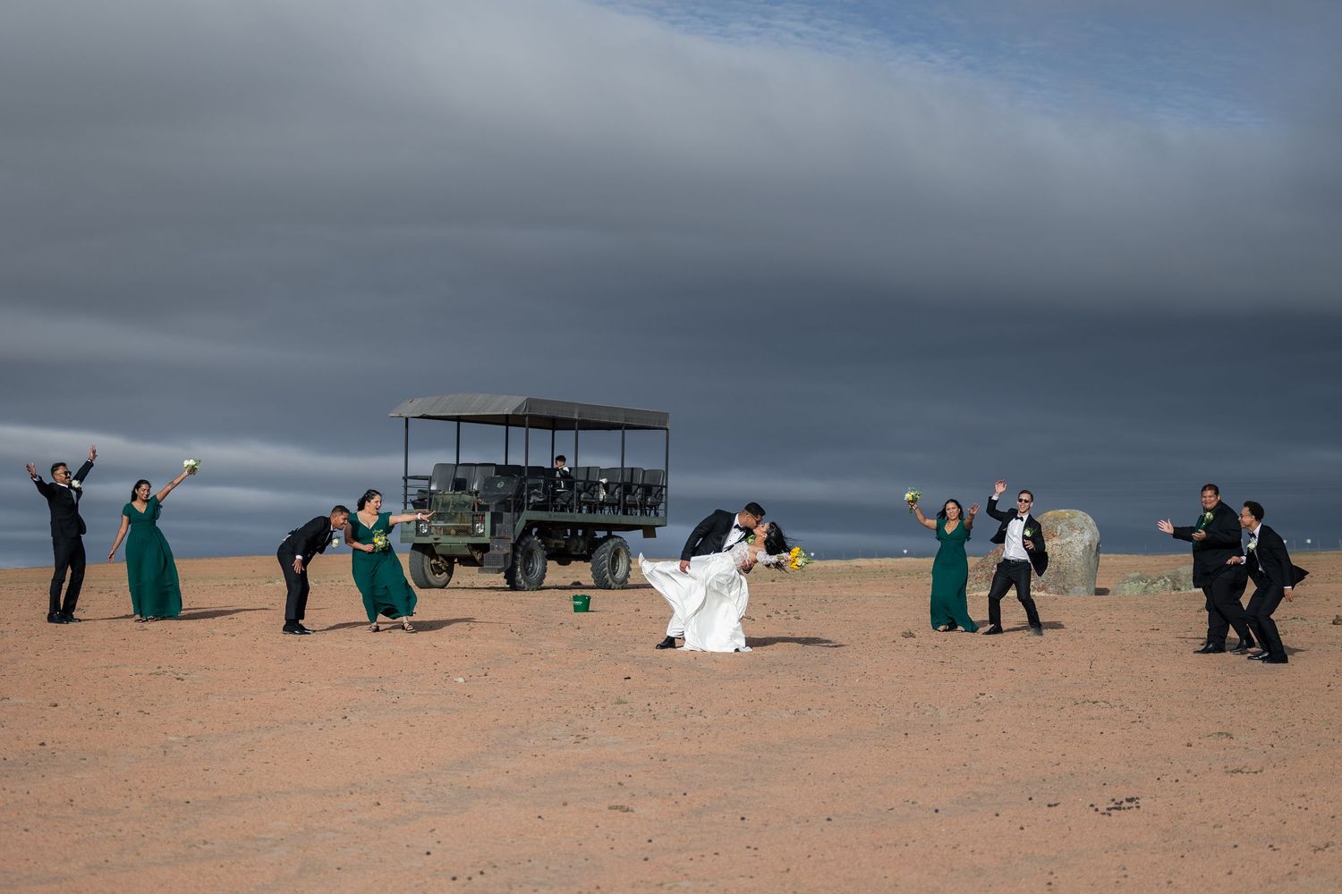 Wedding party celebrating in desert landscape with safari vehicle, bride and groom kissing, bridesmaids in green dresses.