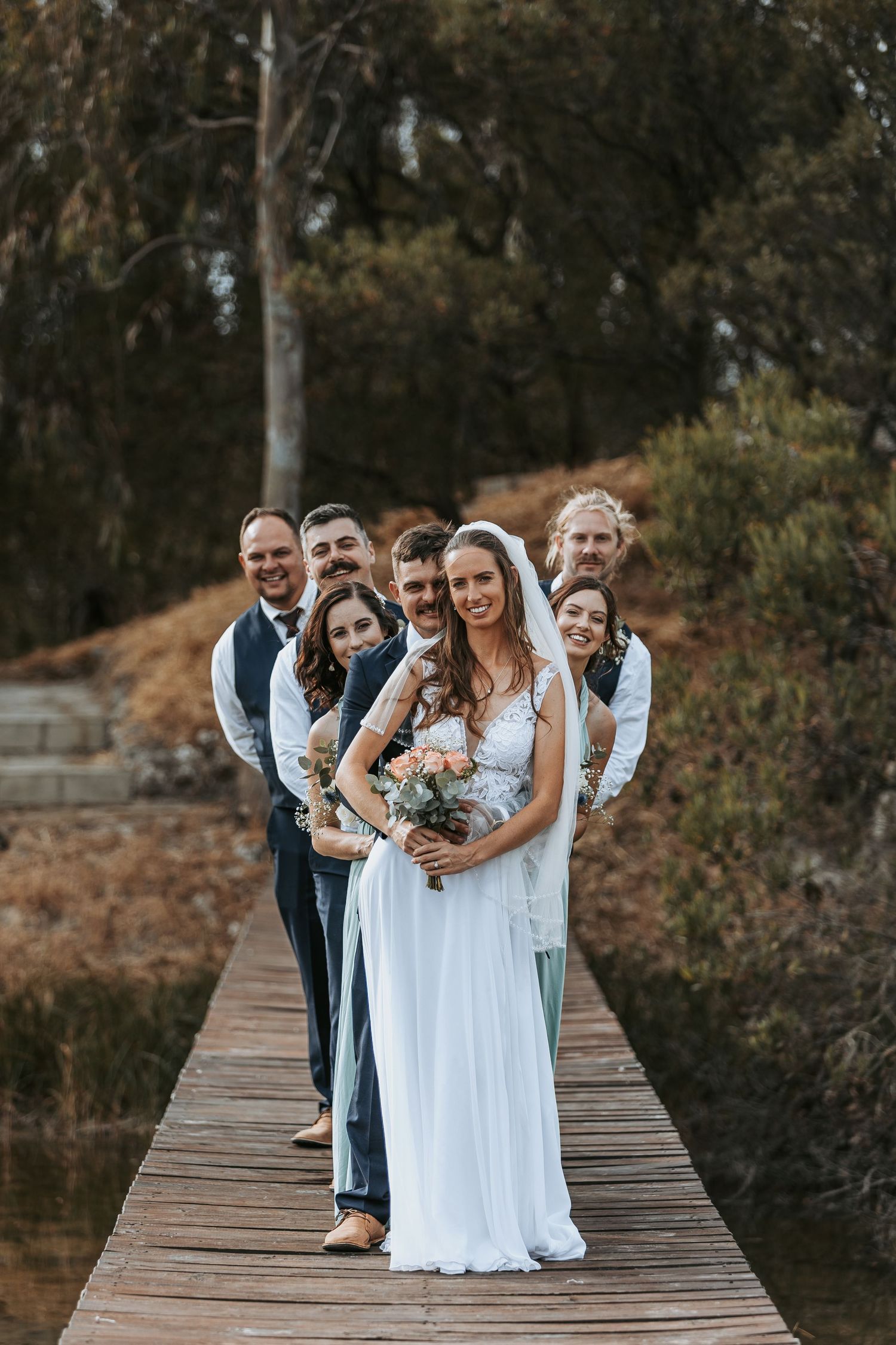 Wedding party laughs together while standing on rustic wooden walkway surrounded by autumn trees.