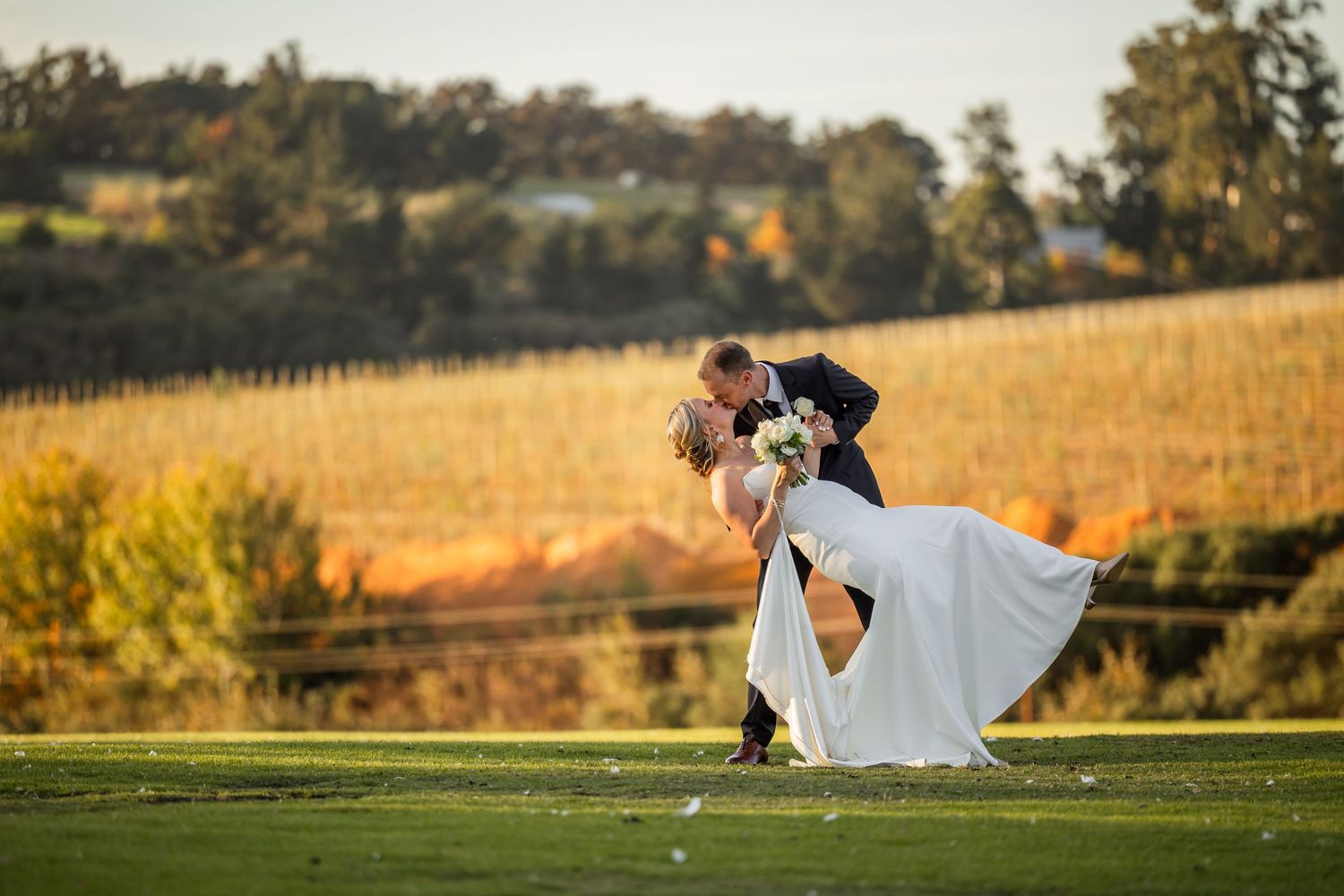 A couple shares a romantic moment in a flowing white dress against a golden sunset in a vineyard setting.