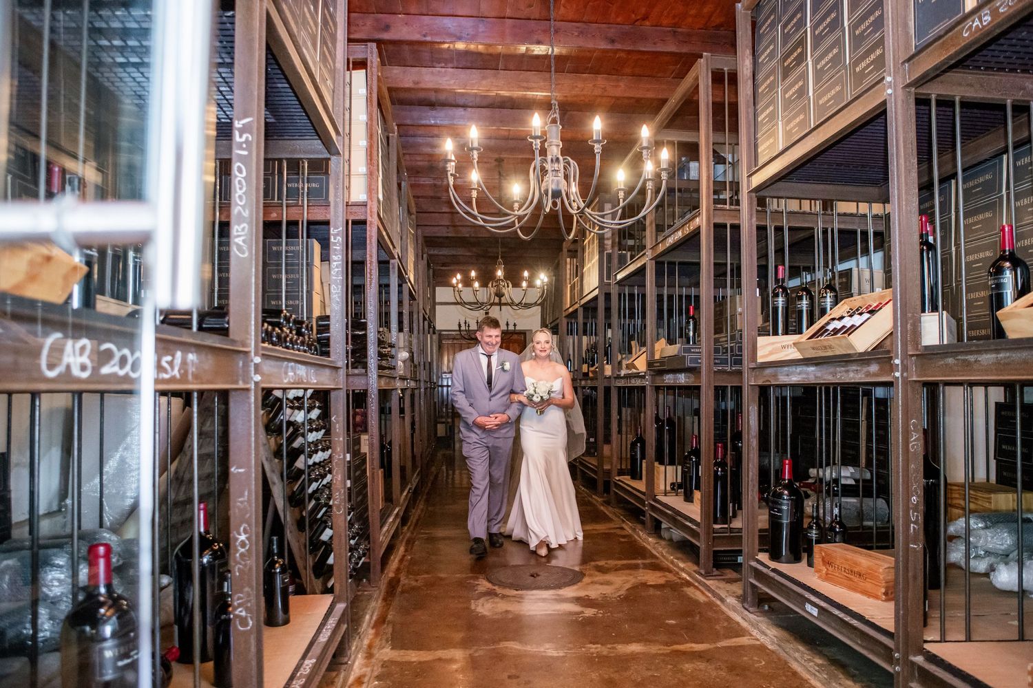 Couple shares romantic moment in elegant wine cellar with wooden racks and chandelier lighting.
