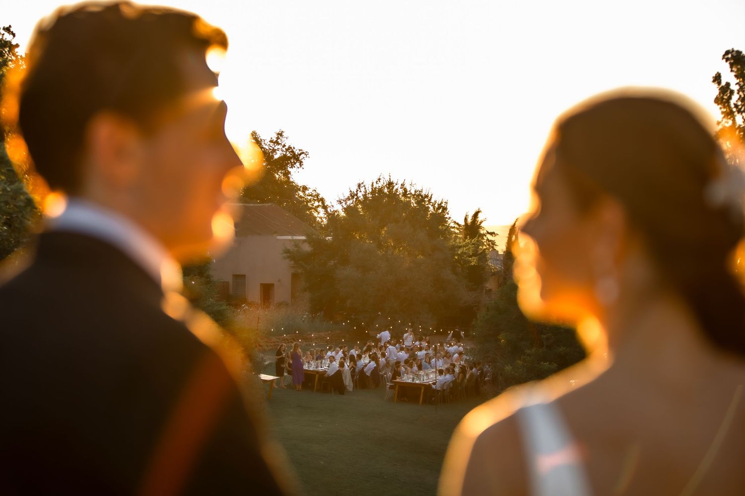 Silhouettes of a wedding couple against a golden sunset with guests visible in the background.