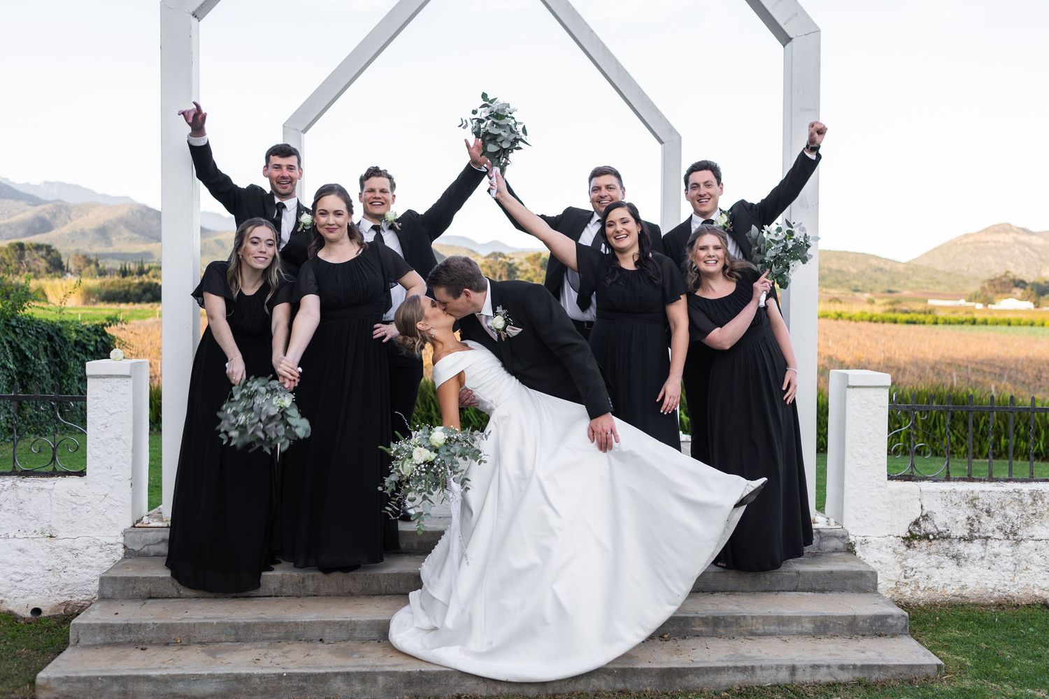 Wedding party kissing and celebrating with raised bouquets under white arch at vineyard venue.