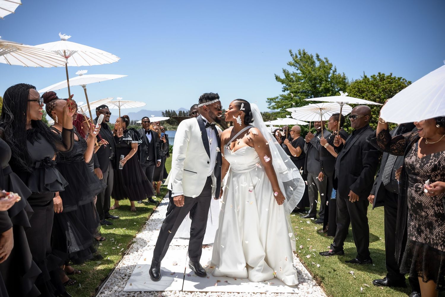 Wedding guests hold white parasols during an outdoor summer ceremony under bright blue skies.