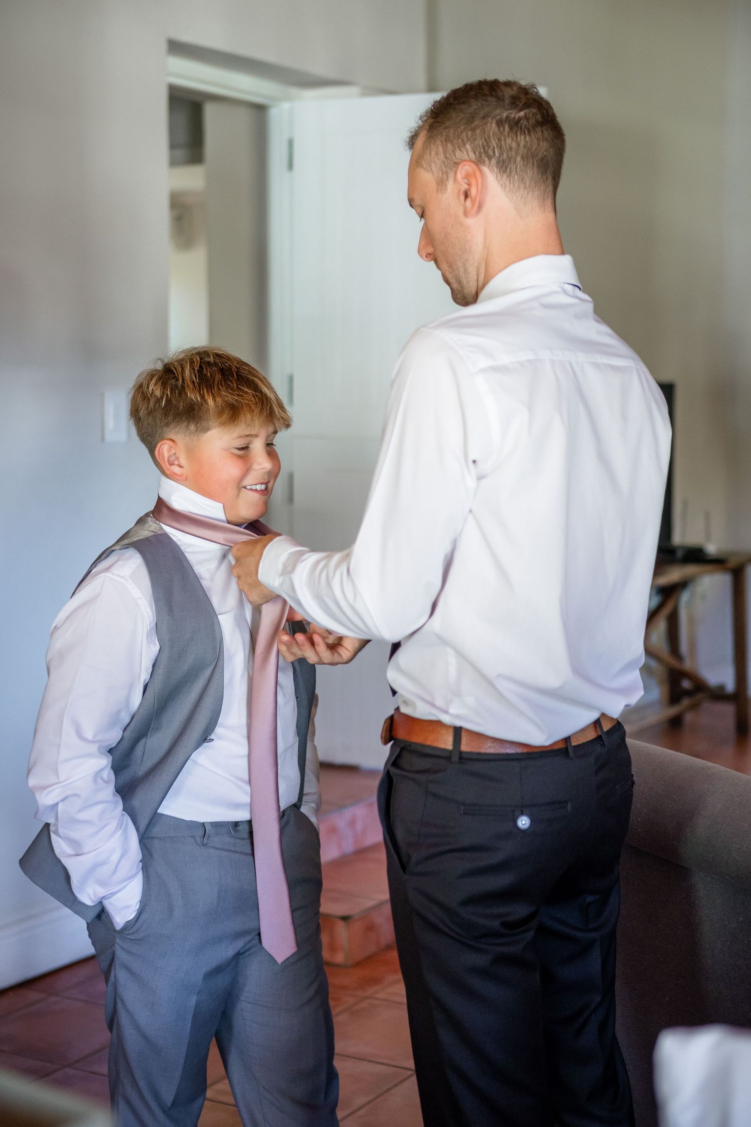 A young boy in a gray vest and pink tie helps adjust a man's white dress shirt.