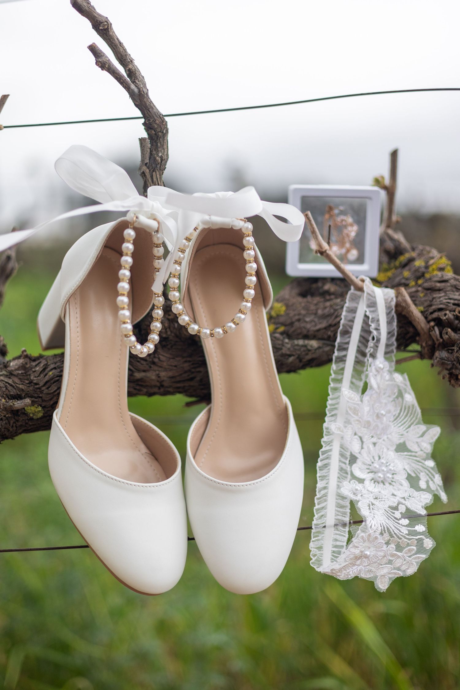 White bridal heels with pearl ankle straps and lace garter hanging on a rustic branch outdoors.