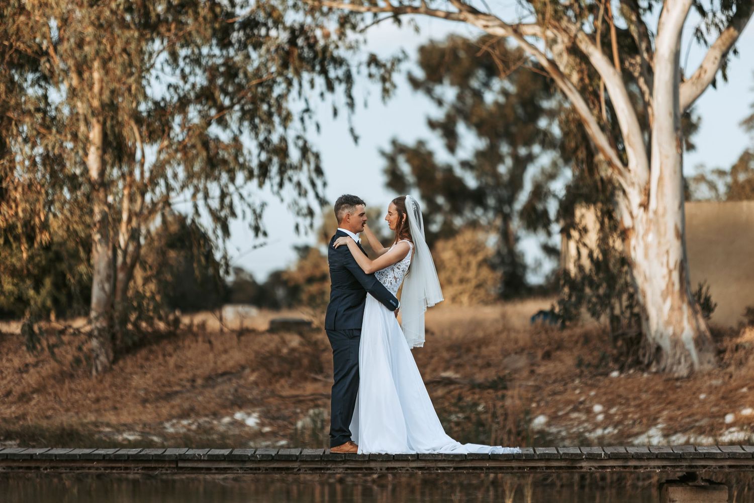 Wedding couple stands together on boardwalk near water with eucalyptus trees in background.
