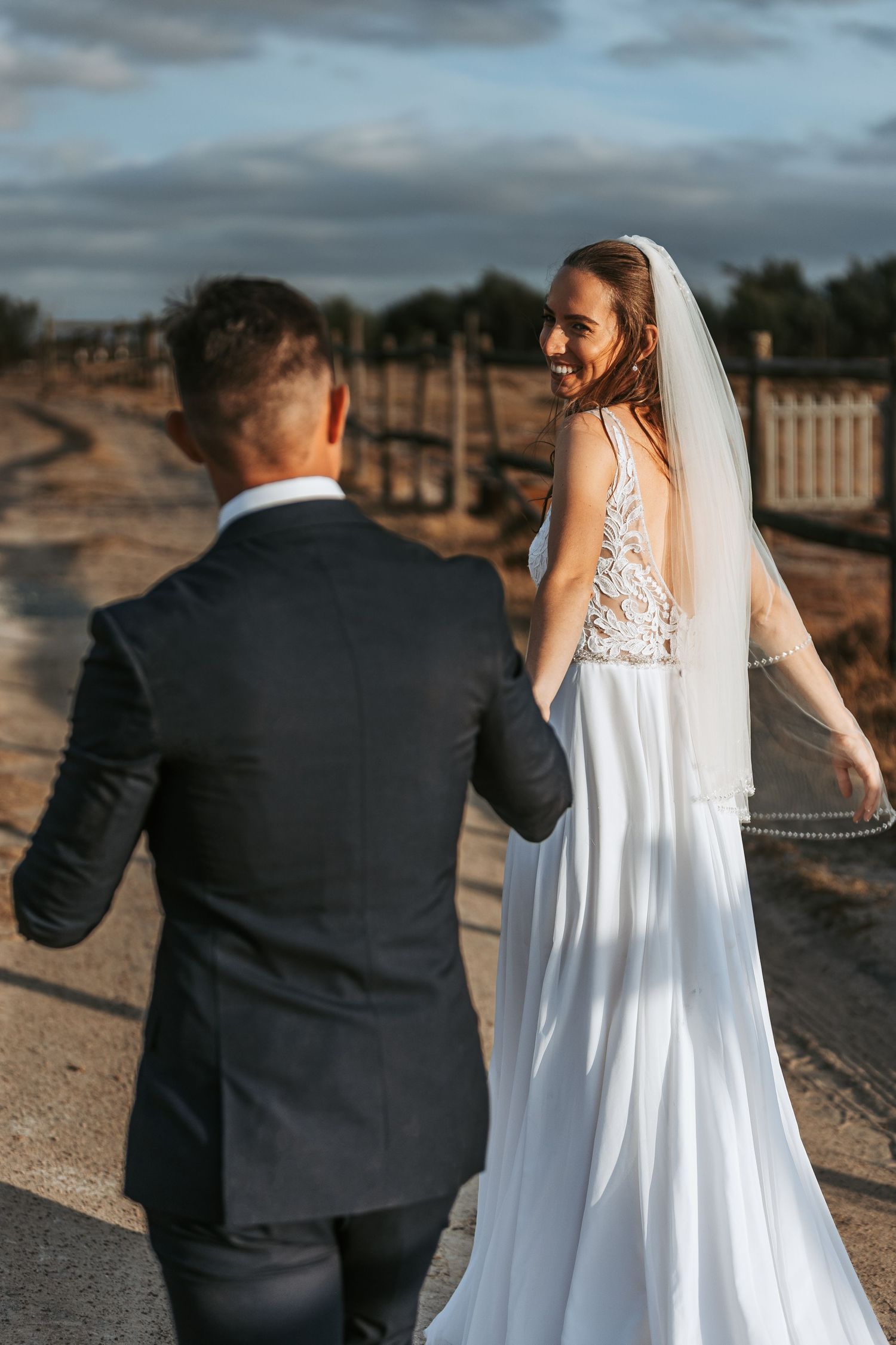 Romantic wedding couple walking along a path at sunset with the bride's white lace dress and veil flowing.