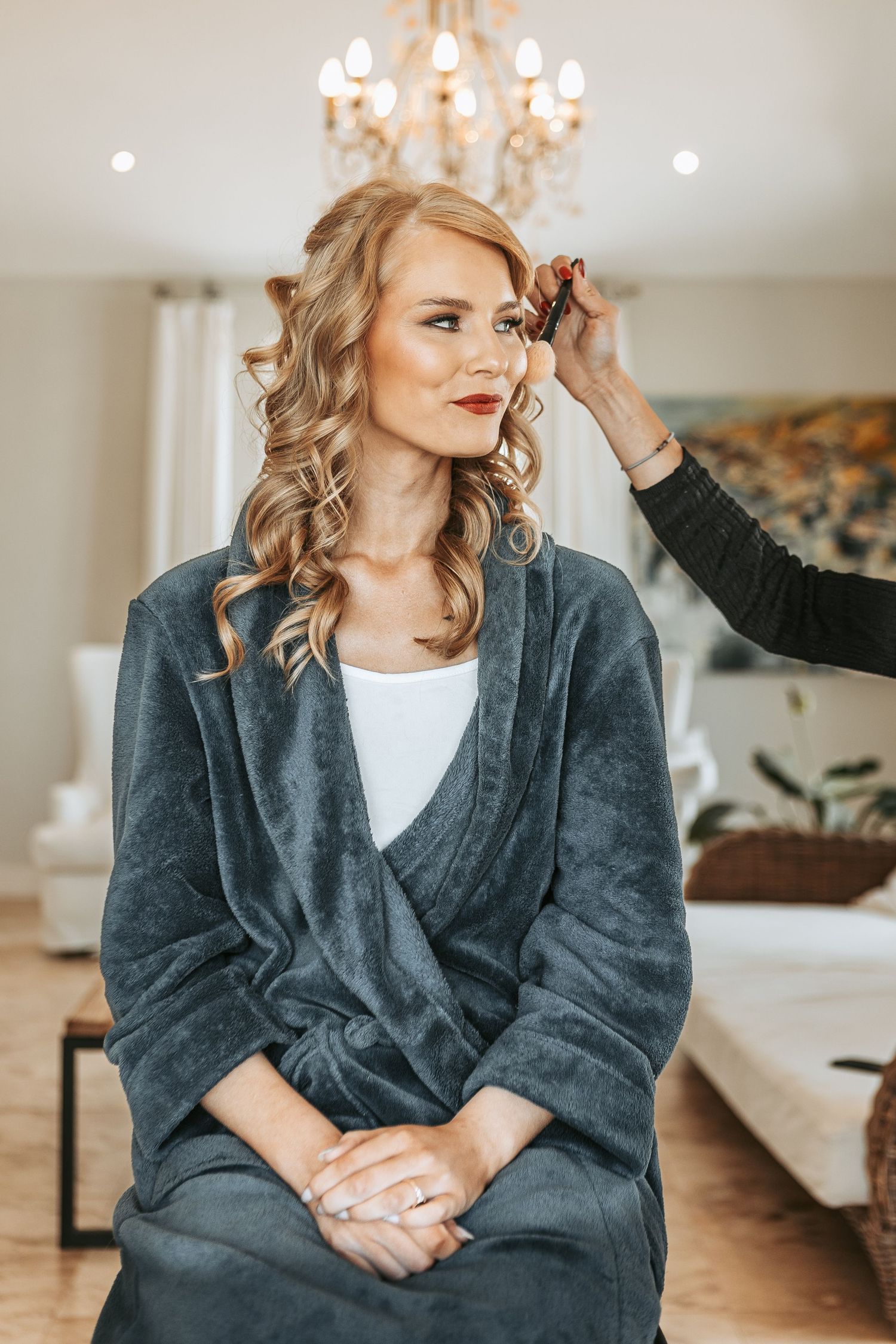 Bride in a plush grey robe getting her hair styled with an elegant chandelier in the background.