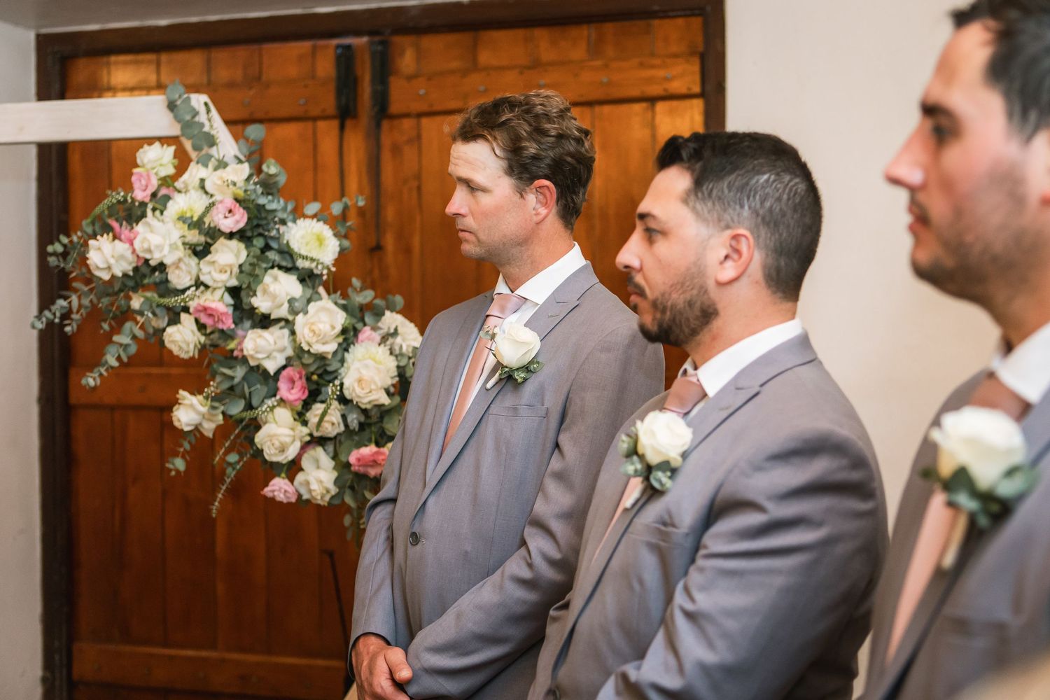 Groomsmen in matching gray suits with pink boutonnieres lined up at wedding ceremony.