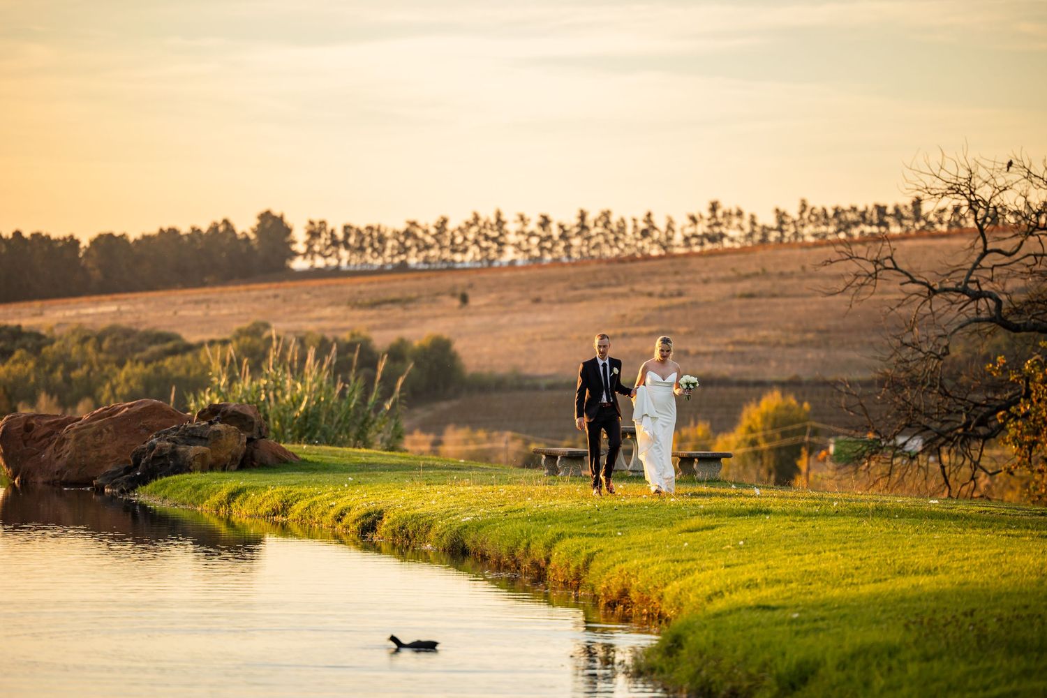 A wedding couple walks along a scenic lakeside path during golden hour with hills and trees in the background.
