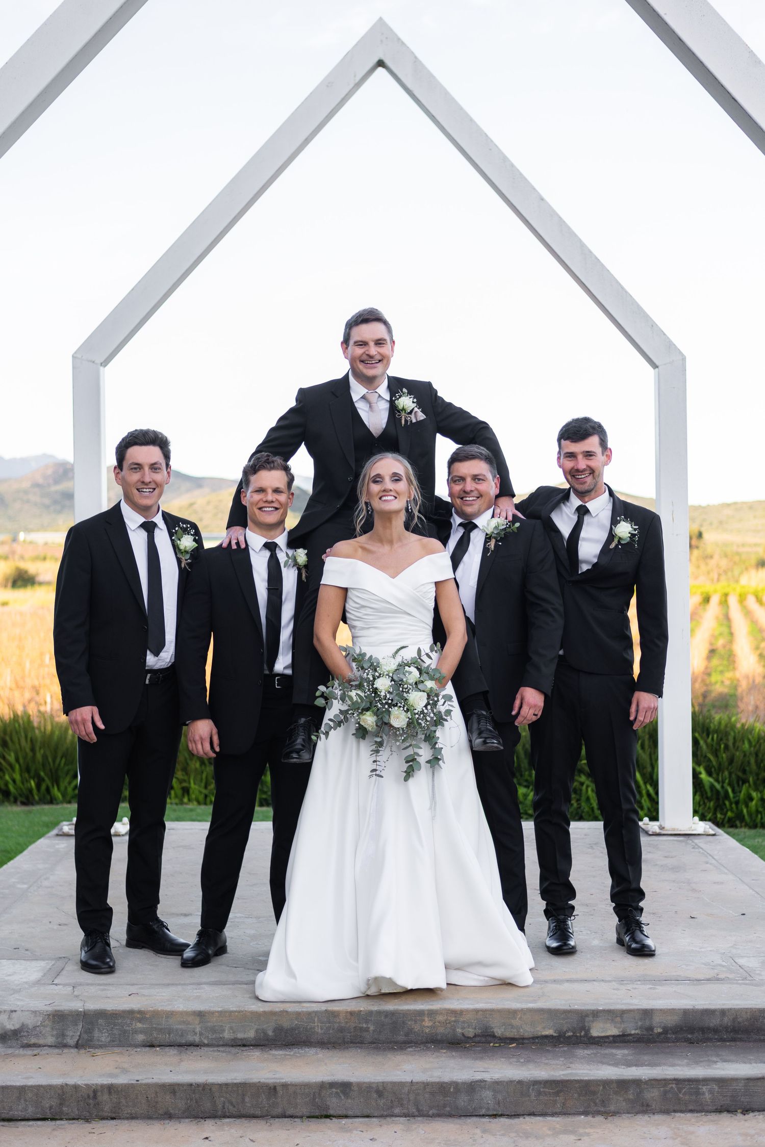 Bride in white off-shoulder gown poses with four groomsmen in black suits before a white arch at a vineyard wedding.