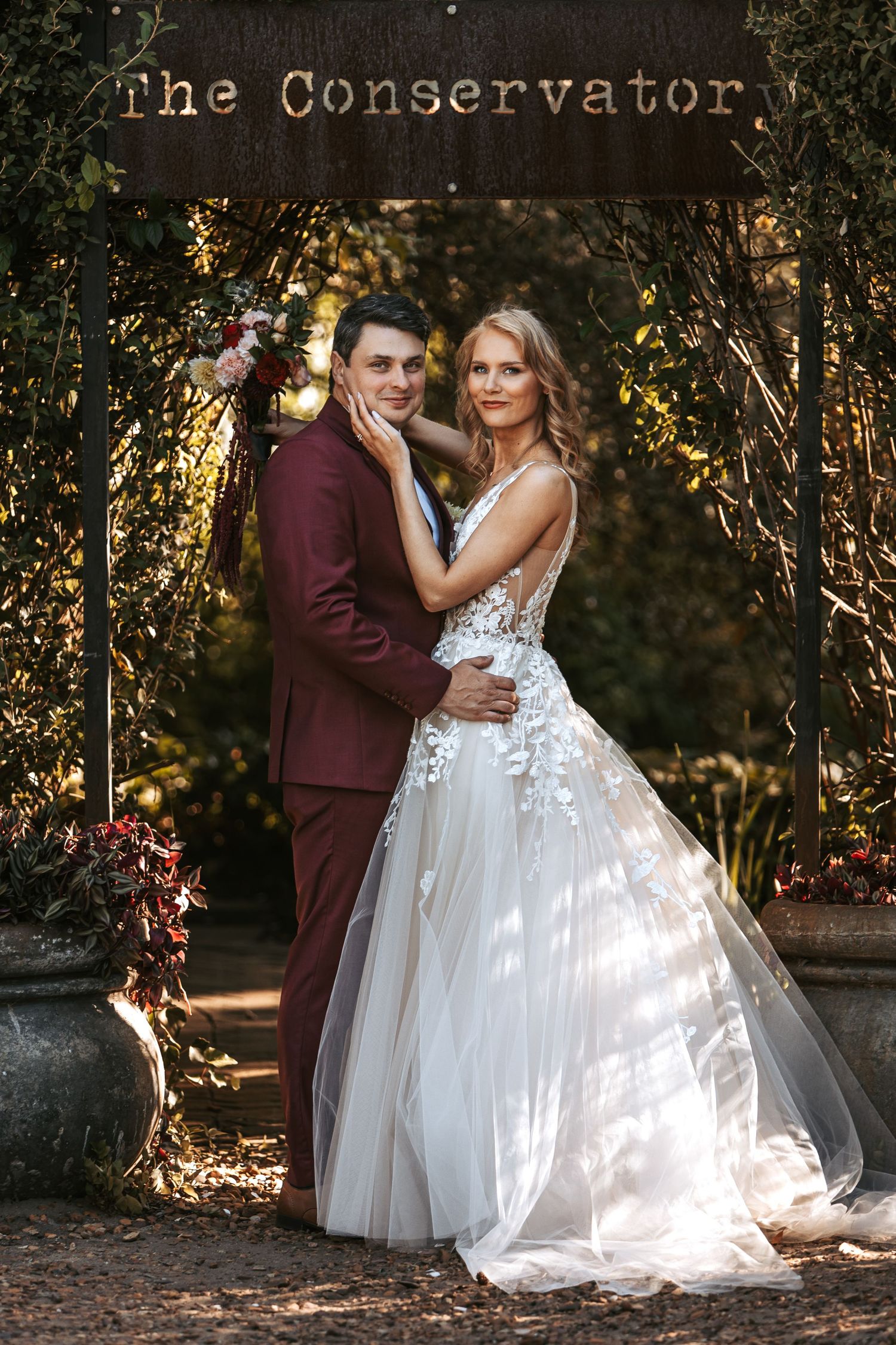 Bride in white lace ballgown and groom in burgundy suit pose under floral arch at The Conservatory wedding venue.