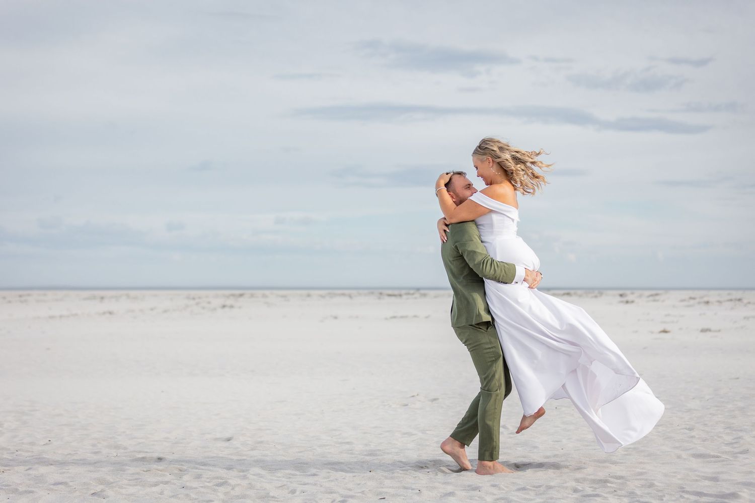 A couple embraces on a serene beach with flowing white dress and green attire against cloudy sky.