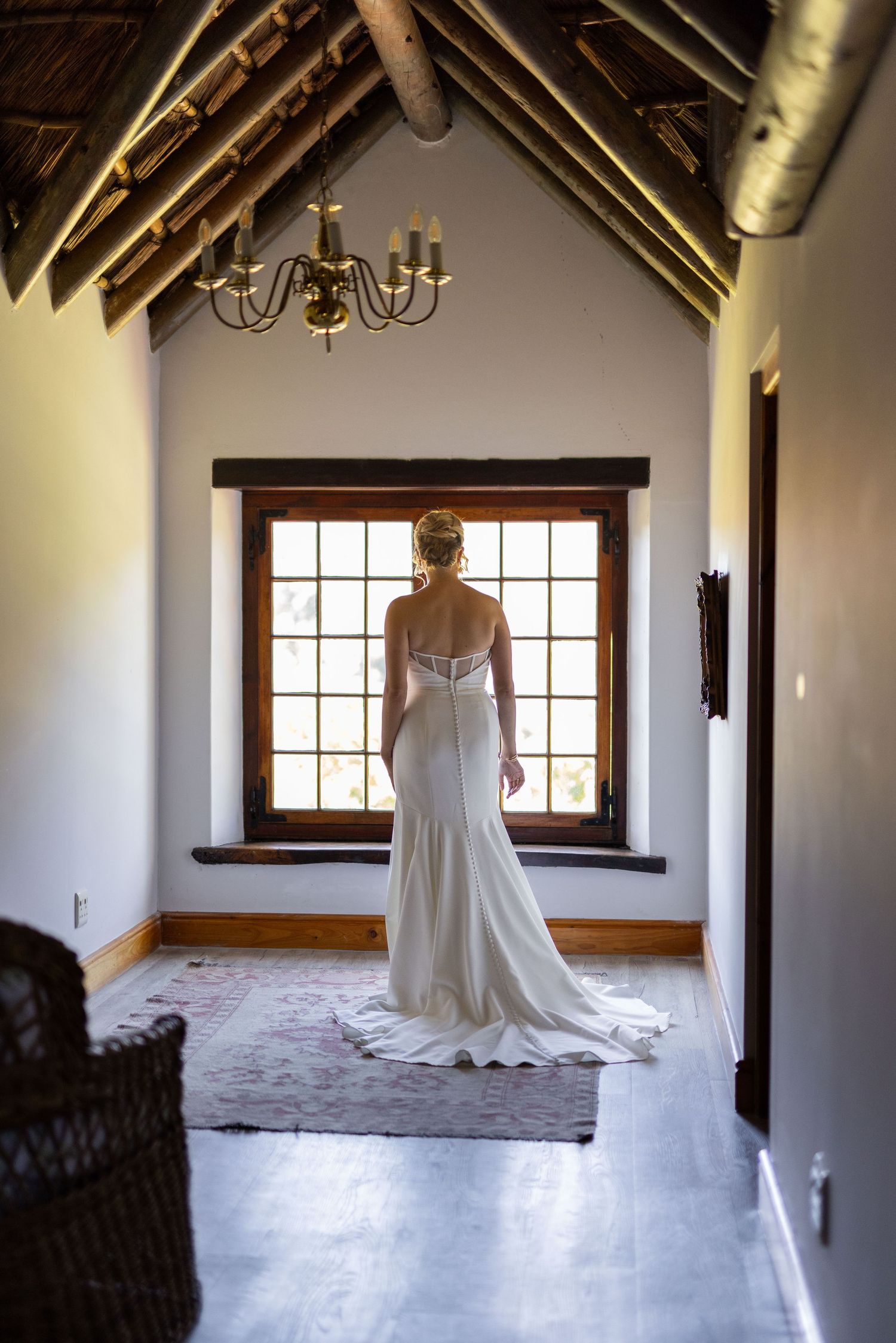 Wedding dress displayed in rustic room with wooden beams and chandelier in natural light.