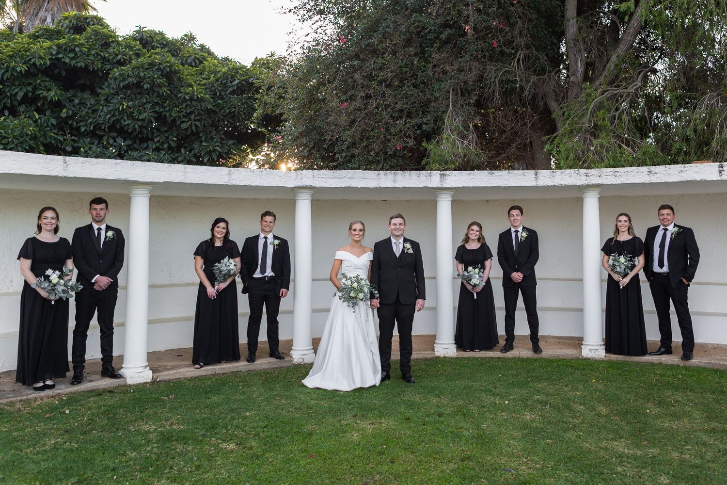 Wedding party in black attire with bride and groom posing at white colonnade.