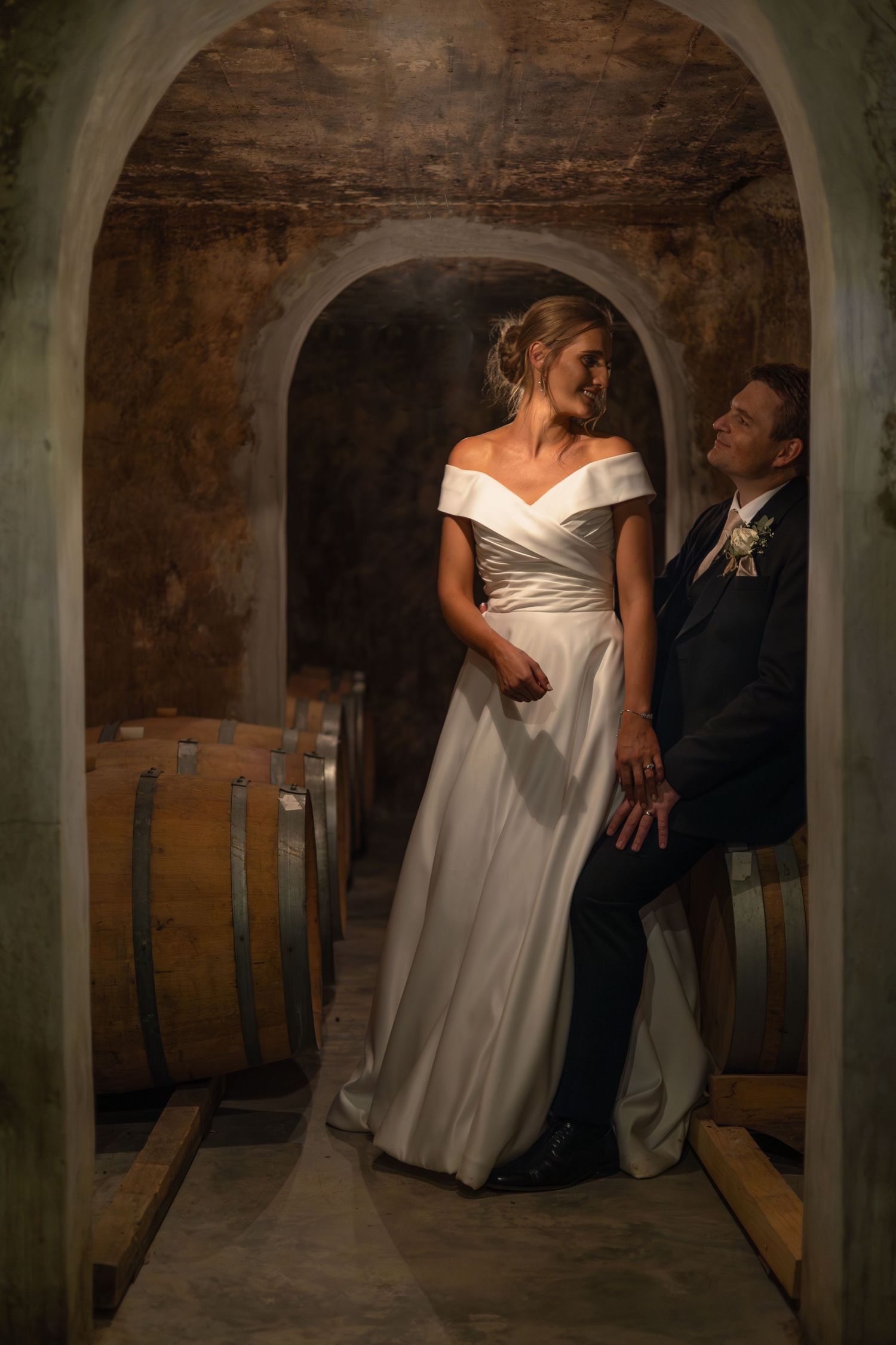 Bride and groom posing romantically in a rustic wine cellar surrounded by wooden barrels under stone arches.