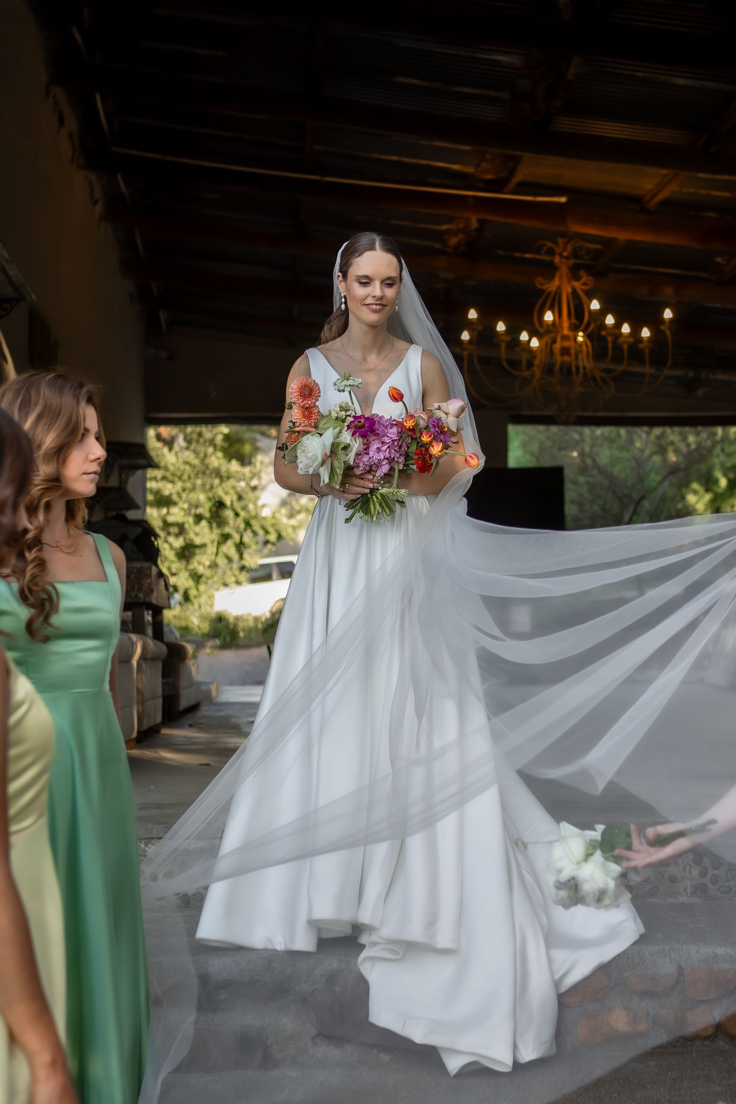 A bride in a flowing white gown holds a vibrant bouquet while attendants in colorful dresses assist with her veil.