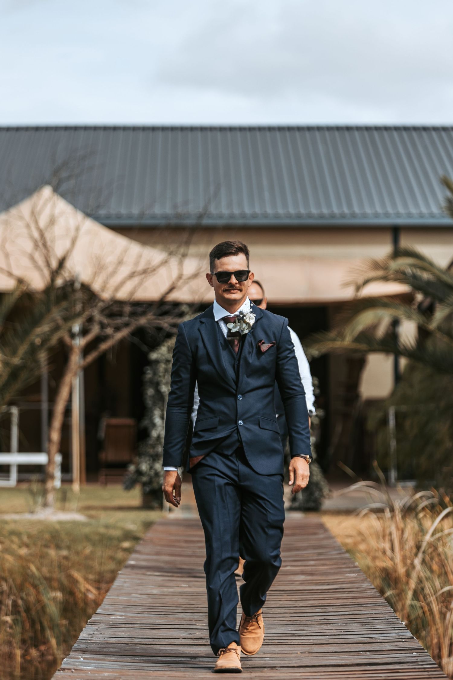 Groom in navy suit walks confidently down wooden boardwalk path during wedding day.