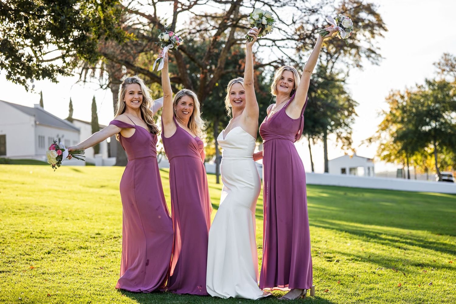Wedding party in purple dresses and white gown posing together in late afternoon sunlight.