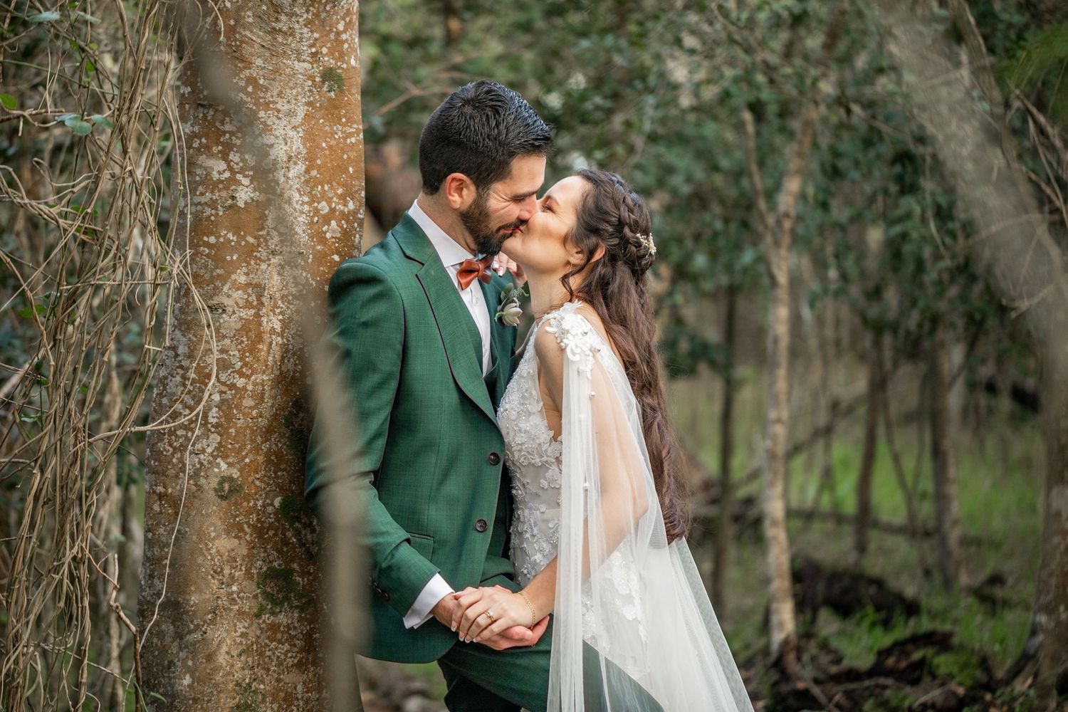 A couple in wedding attire share intimate moments in a wooded outdoor setting with natural greenery.