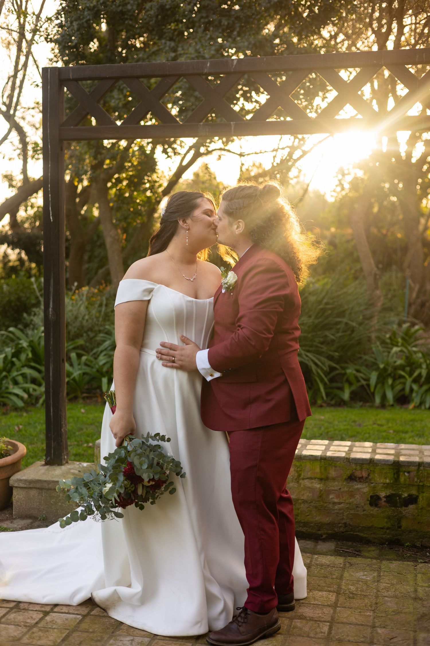 Same-sex couple sharing a kiss at golden hour wedding ceremony, bride in white gown holding bouquet, partner in burgundy suit.