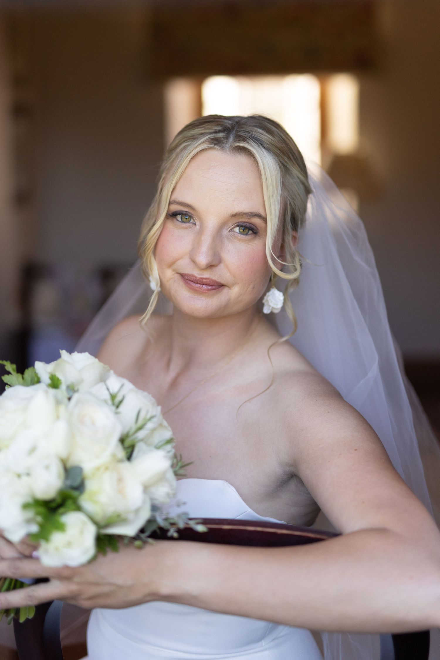 A radiant bride in a white strapless gown holds a bouquet of white flowers while wearing pearl earrings and a veil.
