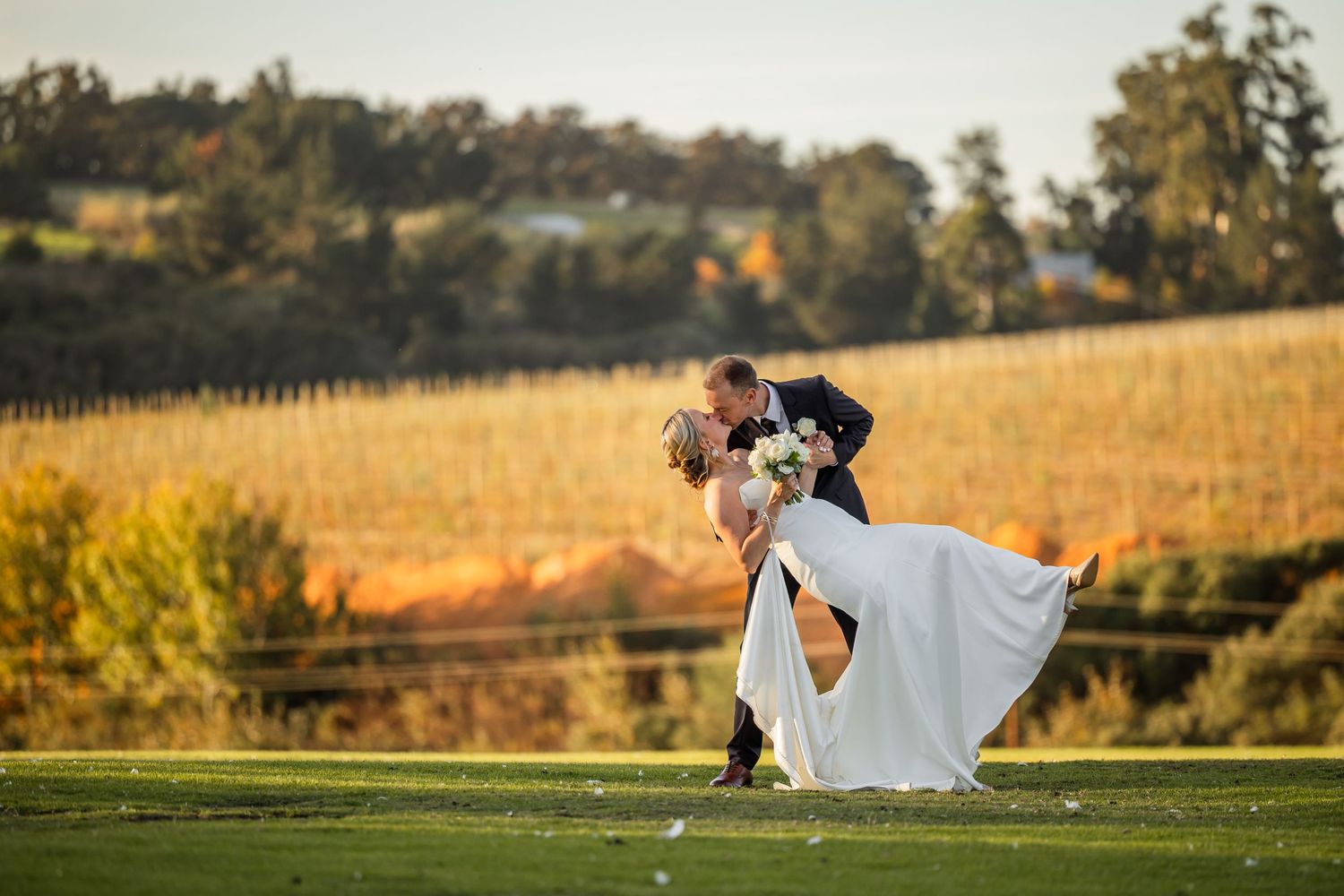 A romantic couple in wedding attire dances together in a scenic vineyard setting during golden hour.