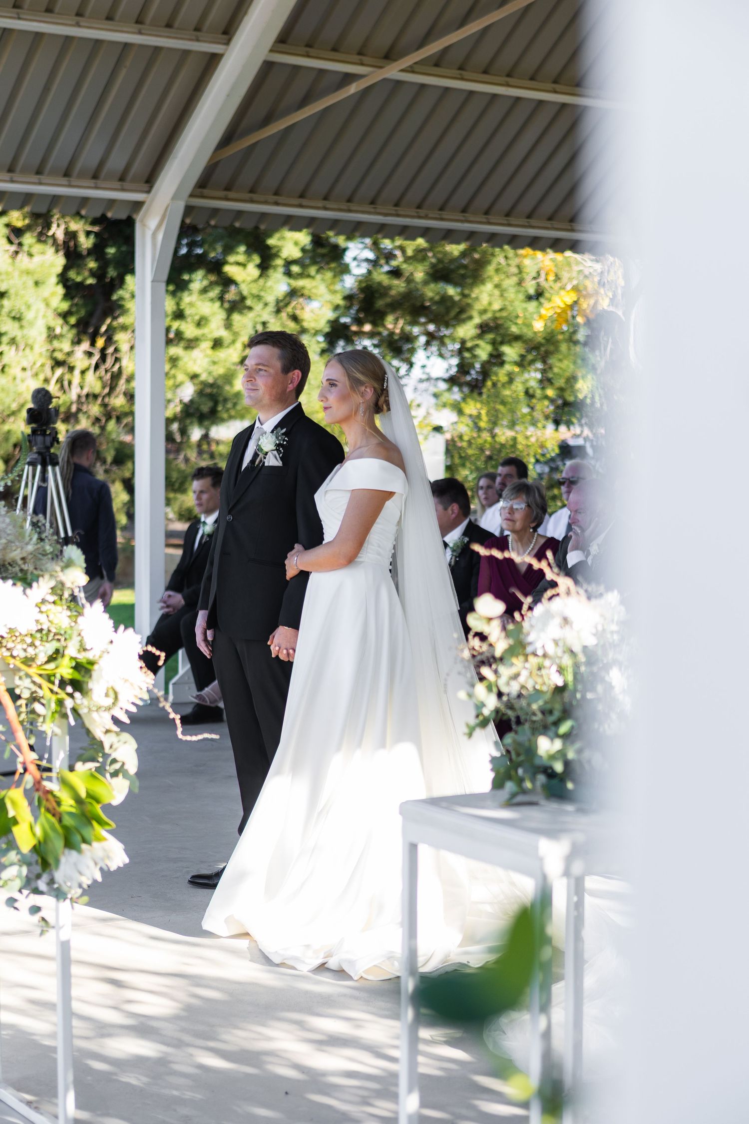 Bride in off-shoulder white gown and groom in black suit walking down aisle at sunlit outdoor wedding ceremony.