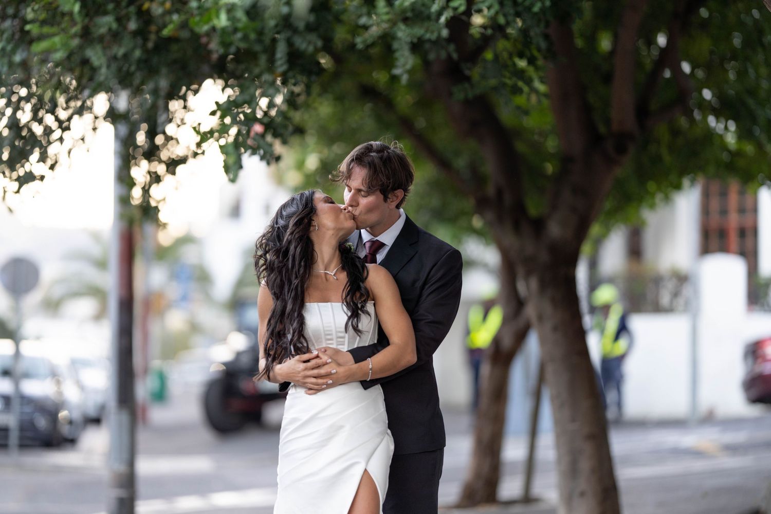 A bride and groom share a romantic kiss outdoors under lush green trees on their wedding day.