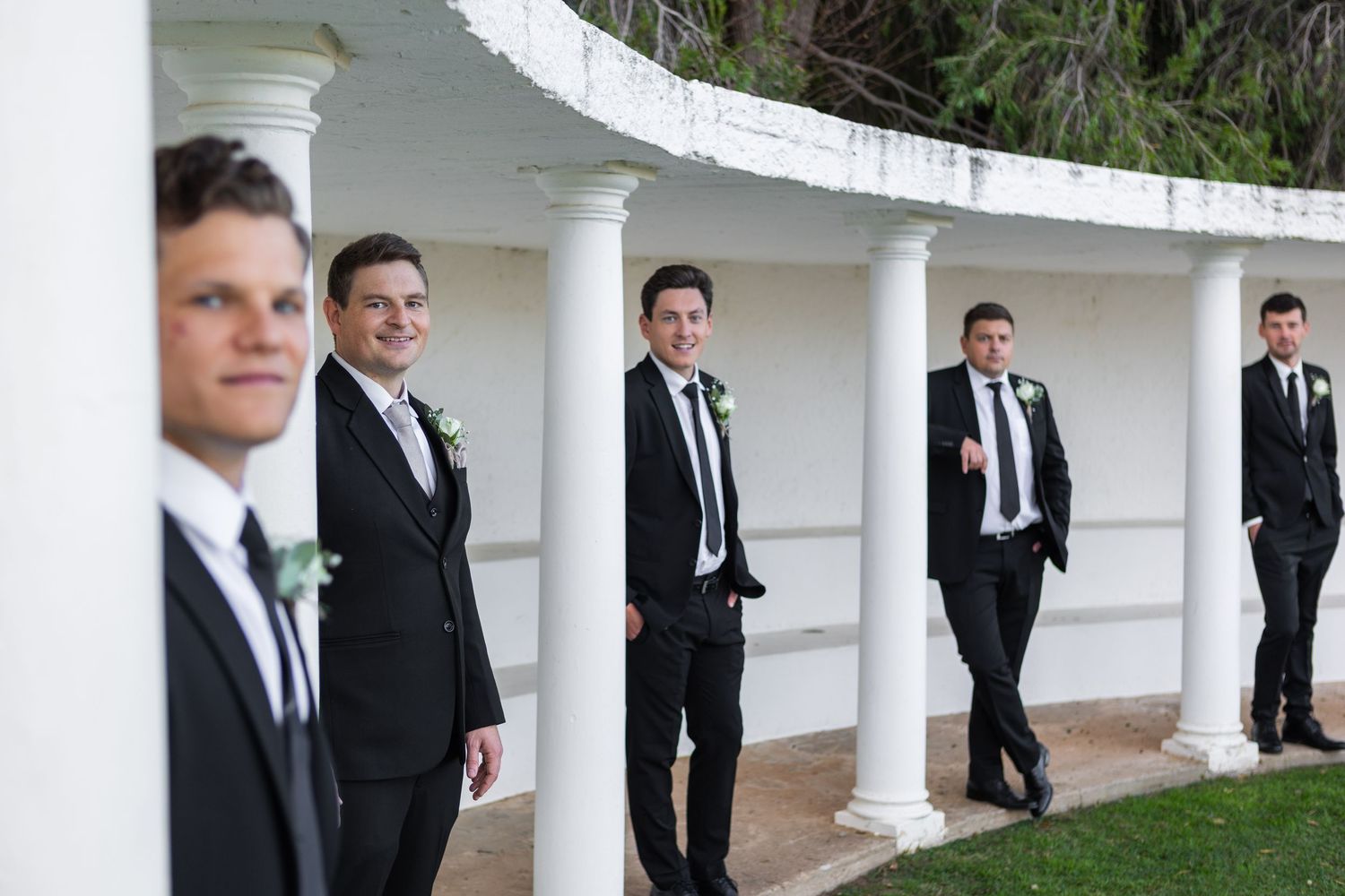 Five groomsmen in black suits casually pose between white columns at an elegant wedding venue.