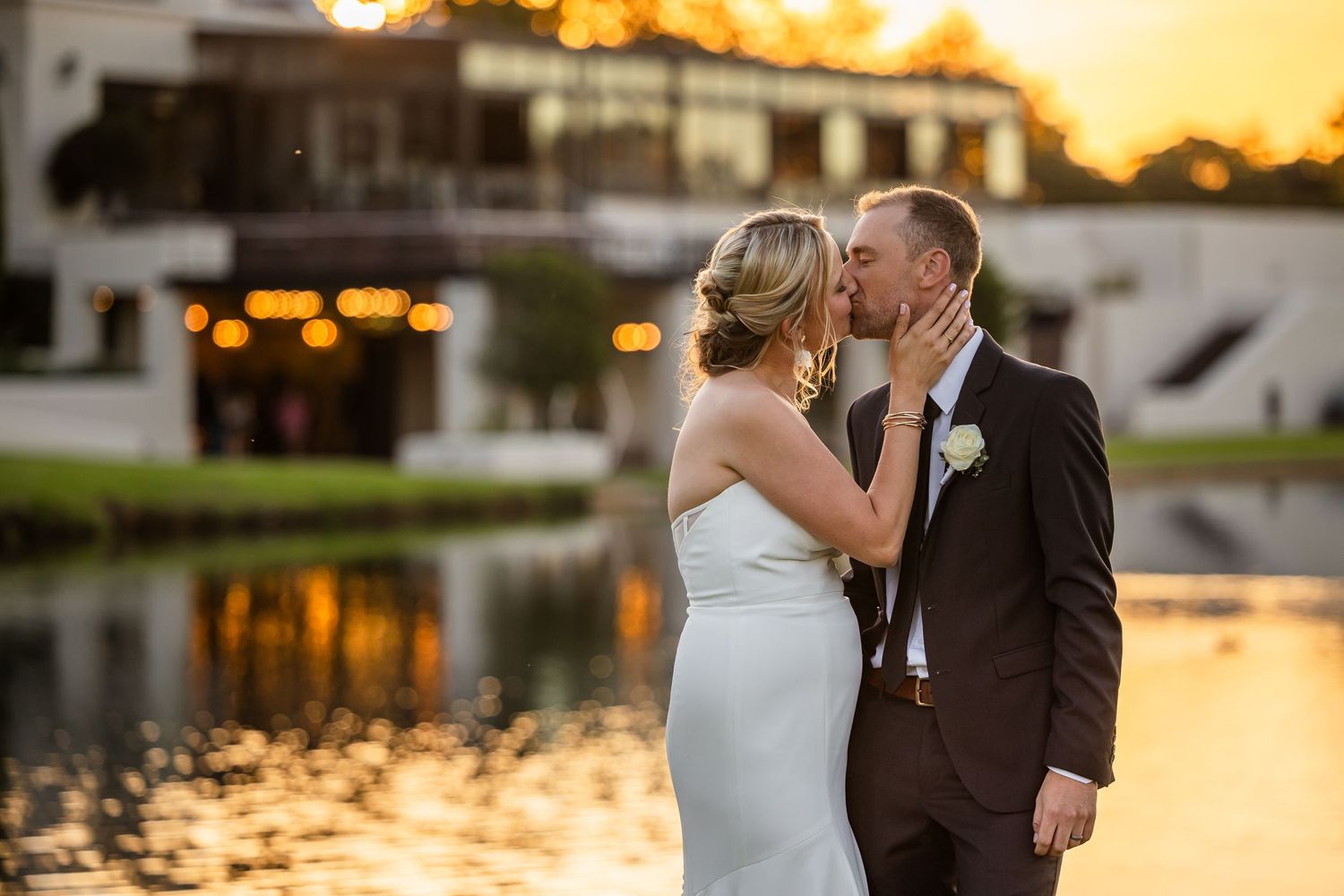 A romantic sunset wedding portrait by a lake with glowing lights in the background.