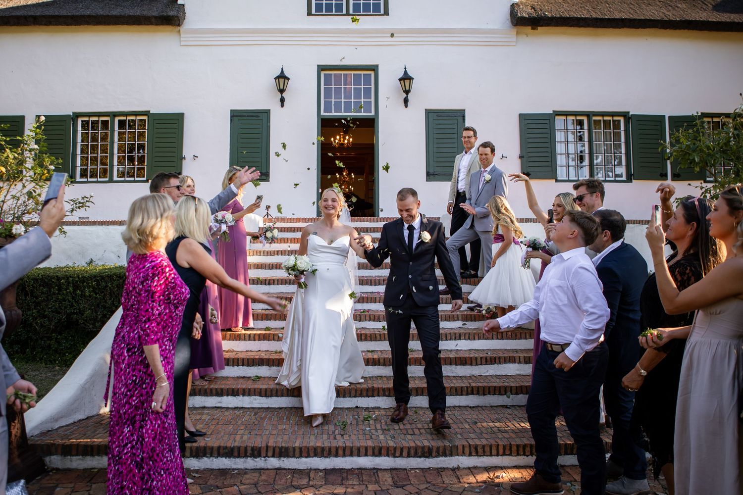 Wedding guests gather on white steps of a colonial style building during an outdoor celebration.