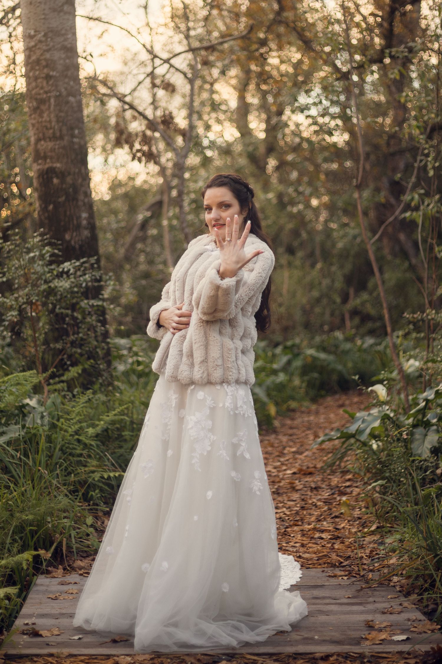 A bride in a flowing white lace gown poses gracefully on a woodland path during golden hour.