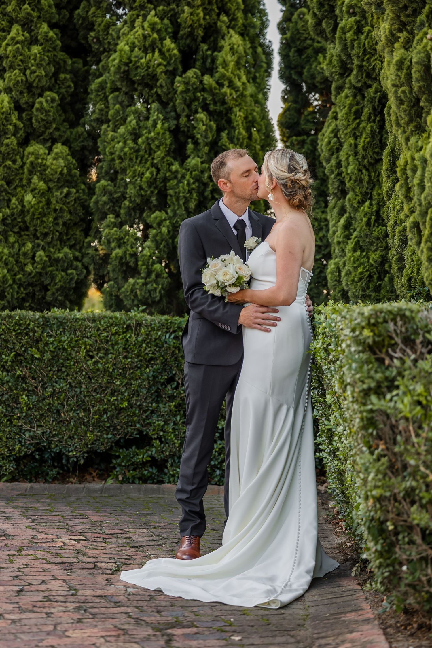 Wedding couple sharing intimate moments on a brick patio with tall cypress trees in the background.