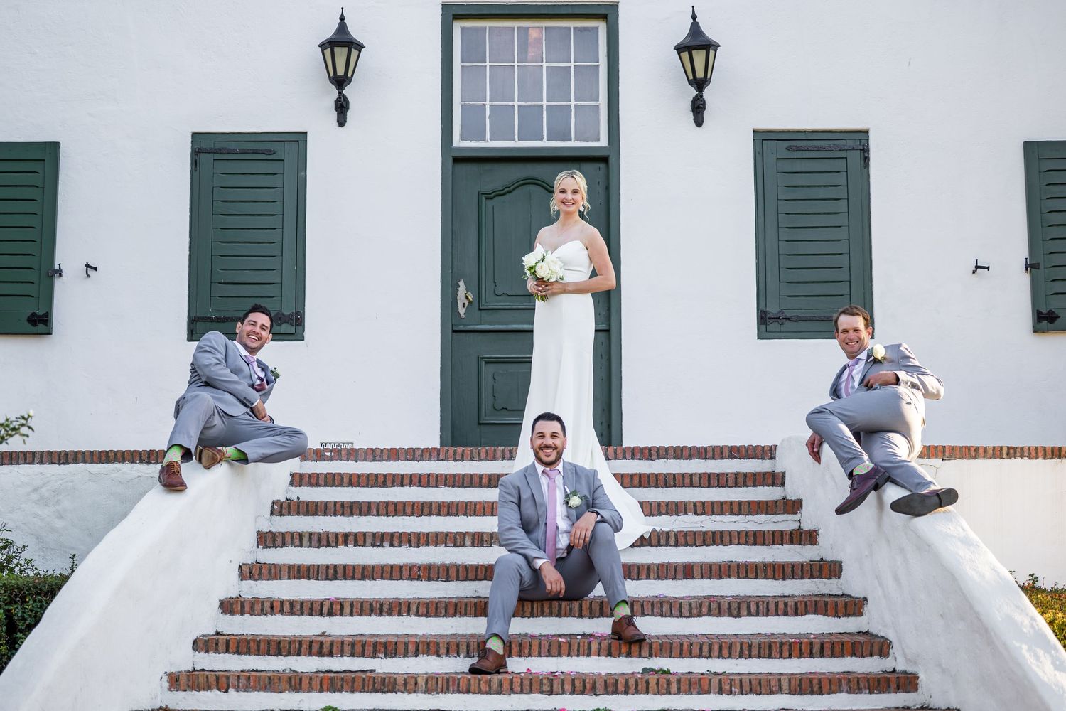 Playful groomsmen pose on white steps with green shutters in sequence of casual shots.
