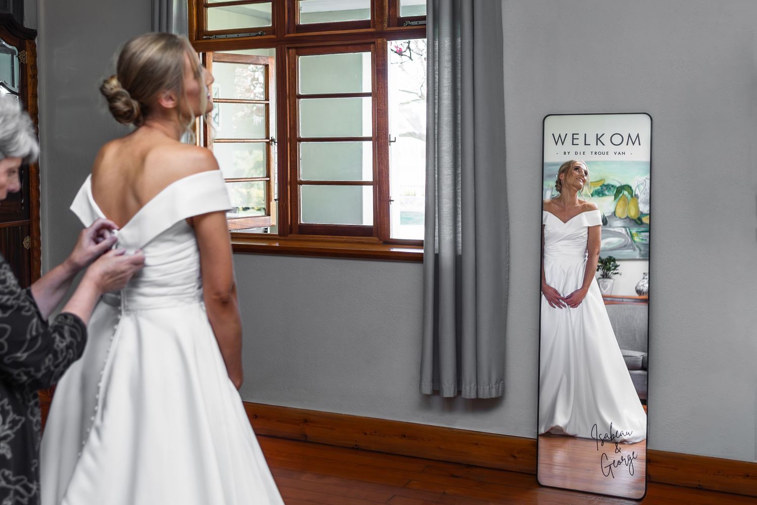 Bride in off-shoulder white gown viewing reflection in welcome mirror before wedding.