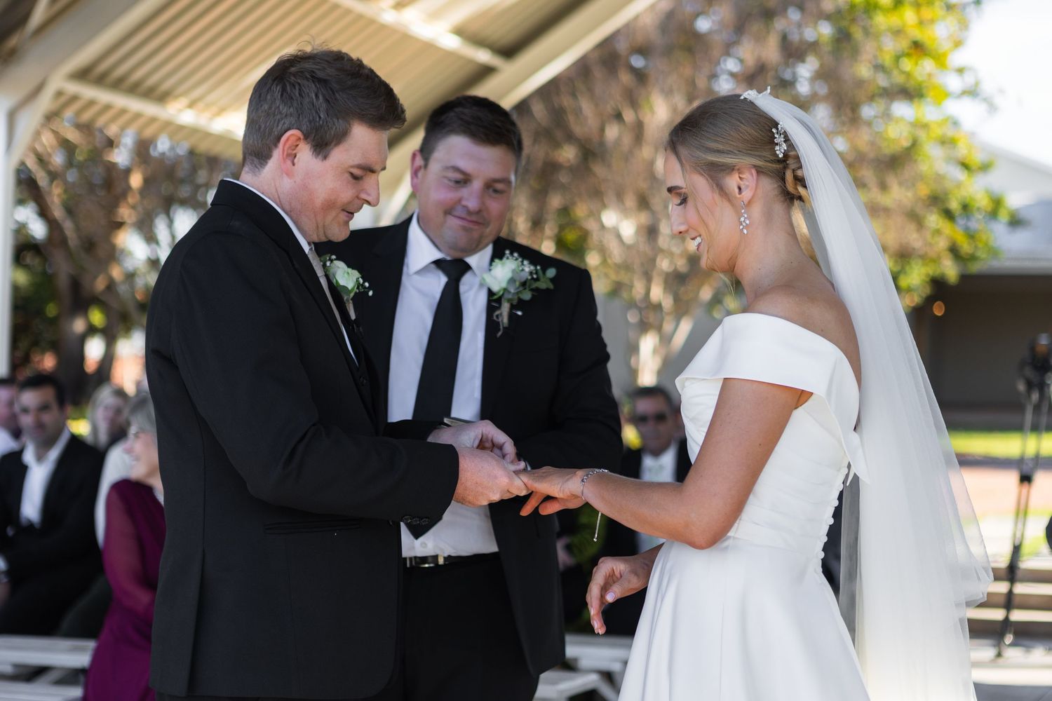 Bride and groom exchanging rings during outdoor wedding ceremony, officiant smiling in background.