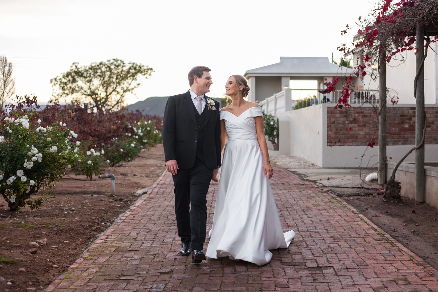Bride and groom walking hand in hand on a brick path, smiling at each other, with roses and a white wall in the background.