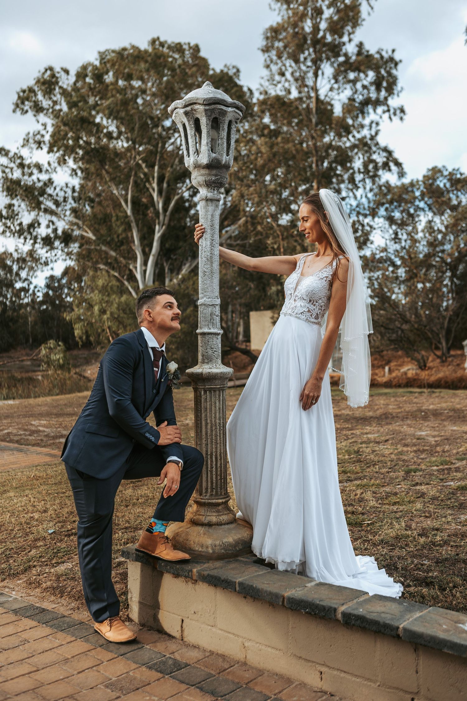 Wedding couple poses playfully on stone wall with vintage lamp posts in outdoor setting.